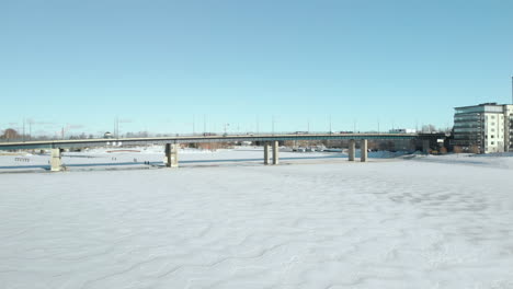 Aéreo,-Disparo-De-Drone,-Sobre-Pielisjoki,-Hacia-El-Tráfico-En-El-Puente-Suvantosilta-Y-Edificios-De-Apartamentos,-En-La-Orilla-Del-Río-En-Un-Día-Soleado-De-Invierno,-En-Joensuu,-Karelia-Del-Norte,-Finlandia