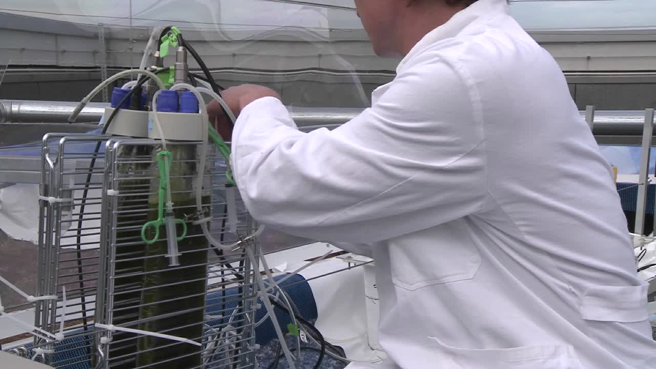 dolly moviéndose de un científico que trabaja con un panel plano hacia otros matraces donde descubrimos a otro científico en el fondo.