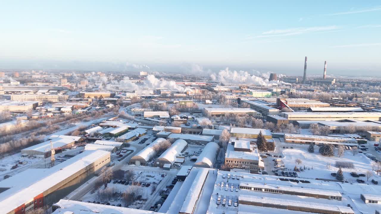 Drone video capturing a vast industrial cityscape blanketed in snow. Smoke billows from chimneys under a clear blue sky, creating a striking winter panorama