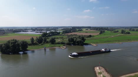 Movimiento-Panorámico-Aéreo-Girando-Alrededor-Y-Siguiendo-Un-Buque-De-Carga-Vacío-Que-Pasa-Por-El-Río-Ijssel-Cerca-De-Zutphen-Con-Llanuras-Aluviales-Y-Turbinas-Eólicas-En-La-Parte-Trasera