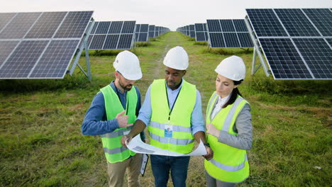 camera zoom out on three multiethnics farm solar engineers talking while looking at blueprint on solar plantation