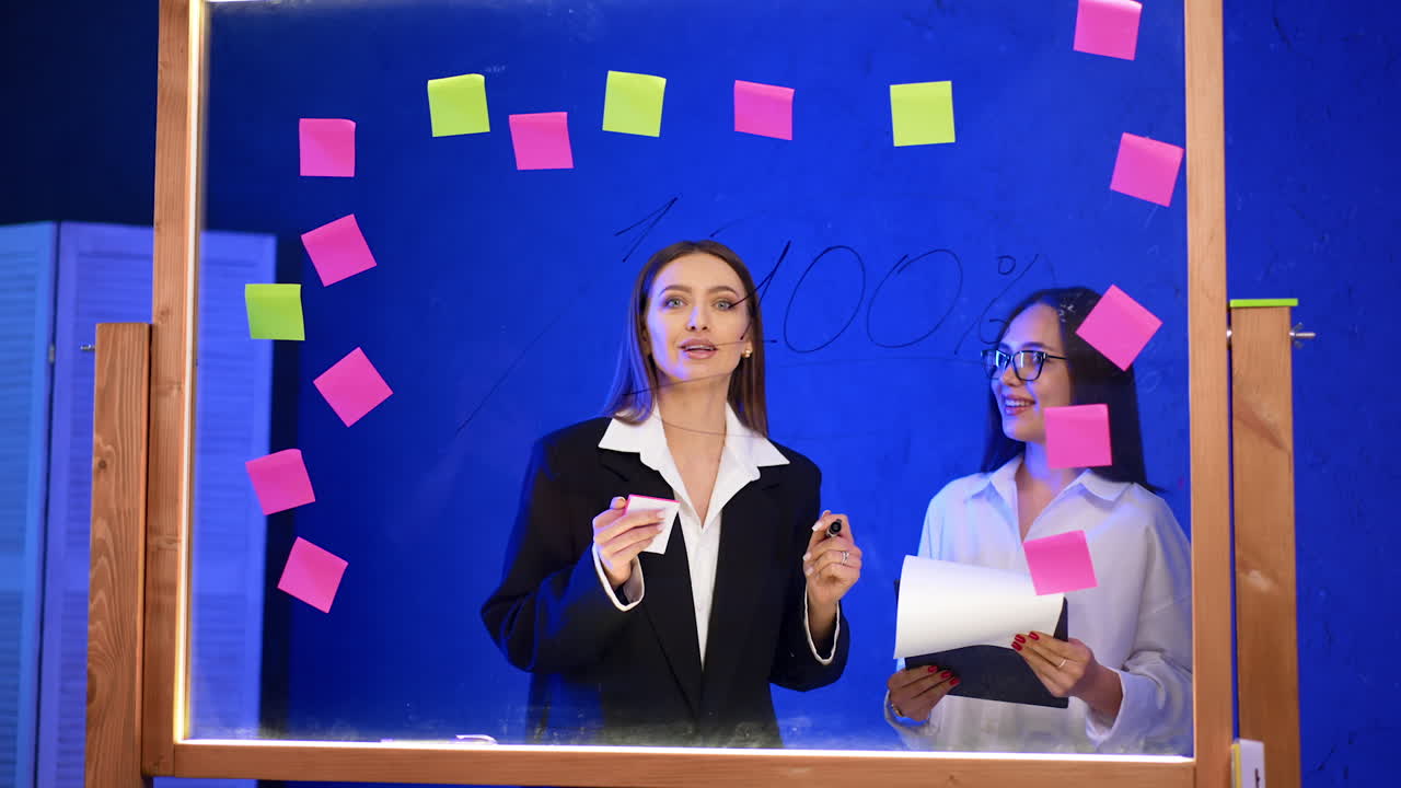 Two ladies stand behind the glass board with stickers on. Woman wearing black jacket talks passionately and gesturing.