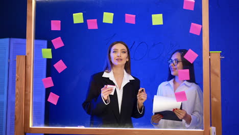 two ladies stand behind the glass board with stickers on. woman wearing black jacket talks passionately and gesturing.