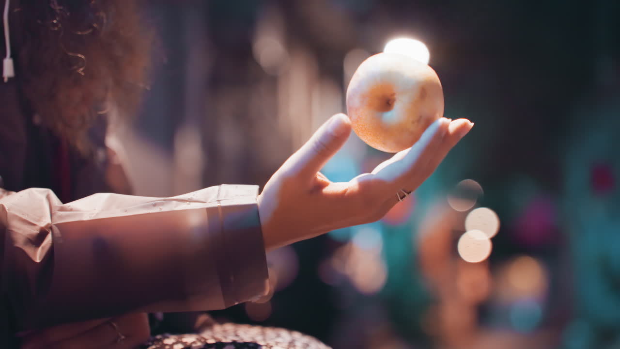 Gros plan d'une main tenant une orange dans une rue nocturne. Une femme aux cheveux bouclés est assise sur un banc, entourée de lumières bokeh chaudes et de reflets de pluie sur le sol. Sa veste est aux couleurs douces, et elle fait un doux geste d'offrande. Scène urbaine intime avec une faible profondeur de champ cinématographique.