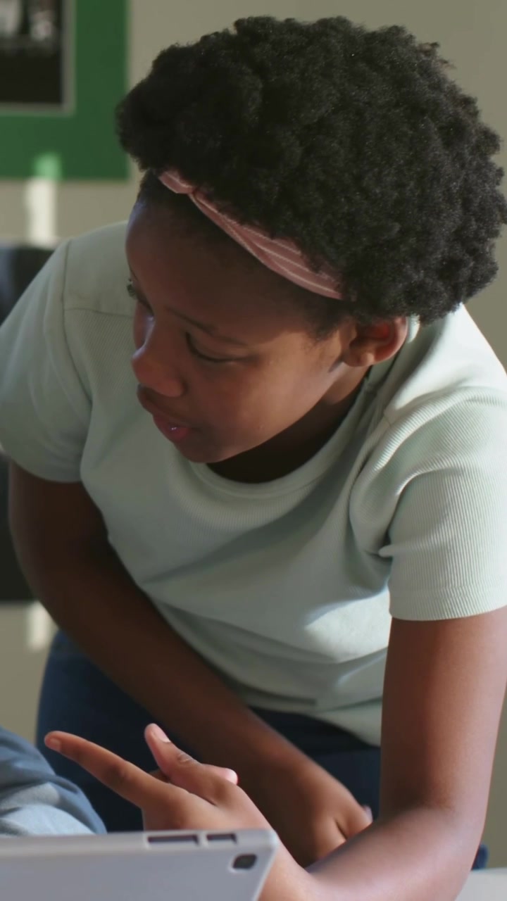 Vertical video: Leaning student in blue top using tablet in class seeing surprise pointing to peer