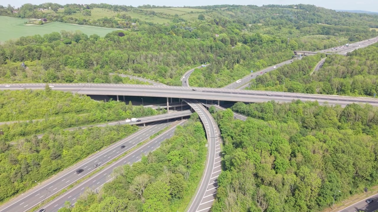 Une perspective aérienne capture des voitures et des camions circulant sur un échangeur autoroutier très fréquenté. Plusieurs routes surélevées traversent des forêts verdoyantes et luxuriantes et des collines ondulantes sous un soleil de jour éclatant