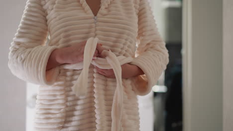 woman with manicure tightens knot on robe in shower on blurred background. person ties plush bathrobe with belt after taking shower slow motion