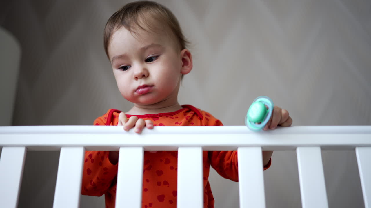 Lovely baby with cute plump cheeks standing in the cot. Adorable little boy holding a pacifier in hand. Low angle view.