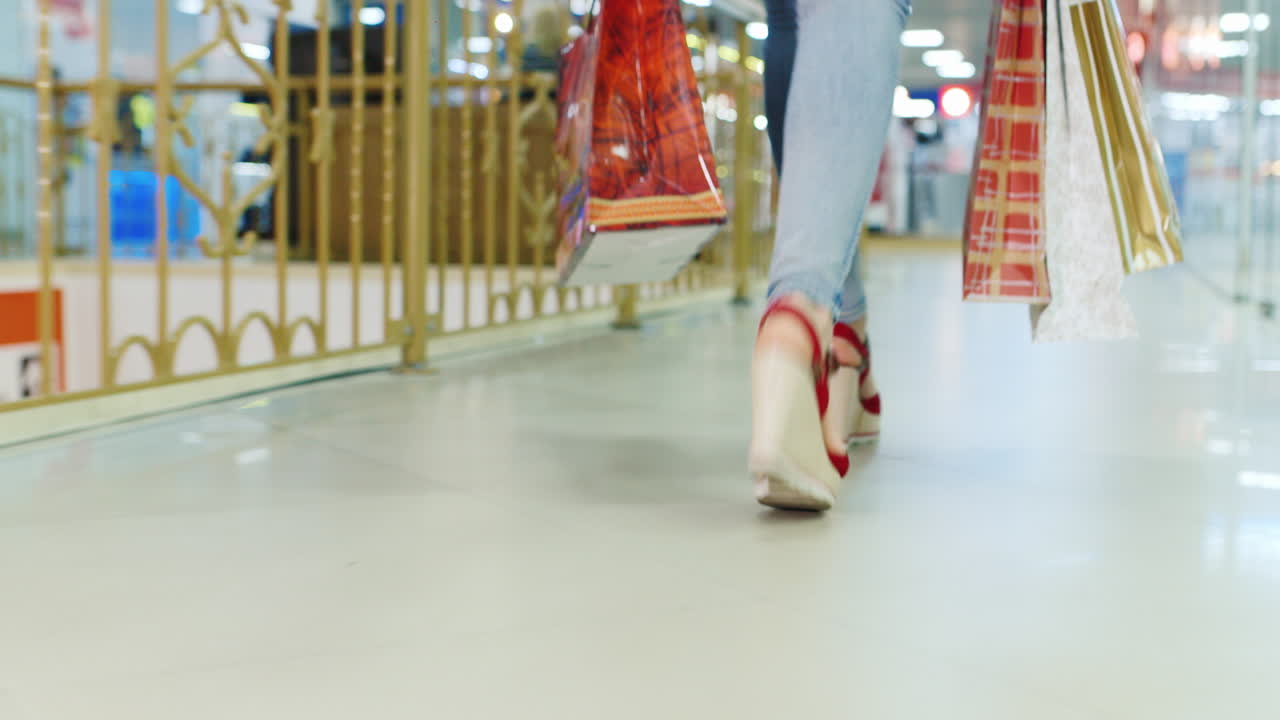 Ung kvinde i jeans og røde sko går i indkøbscentret, set bagfra. Kun benene er synlige i billedet.