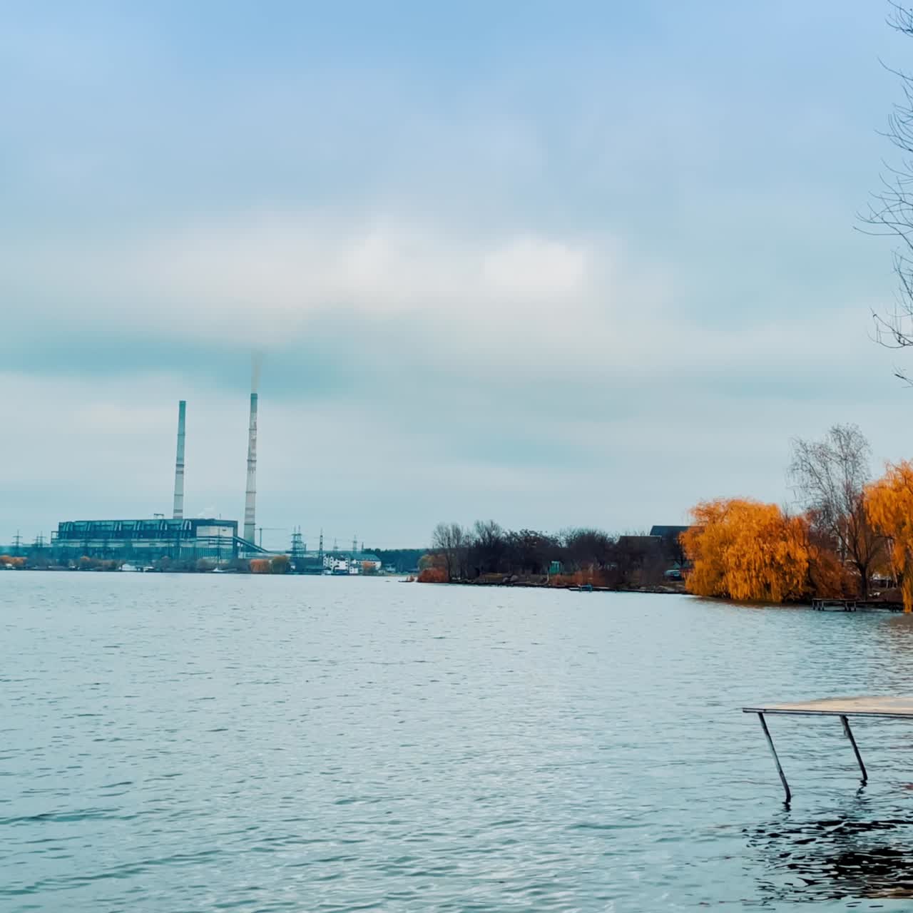 Little footbridge with comfortable deckchair at the river. Yellow weeping willows on the waterfront. Industrial plant at backdrop