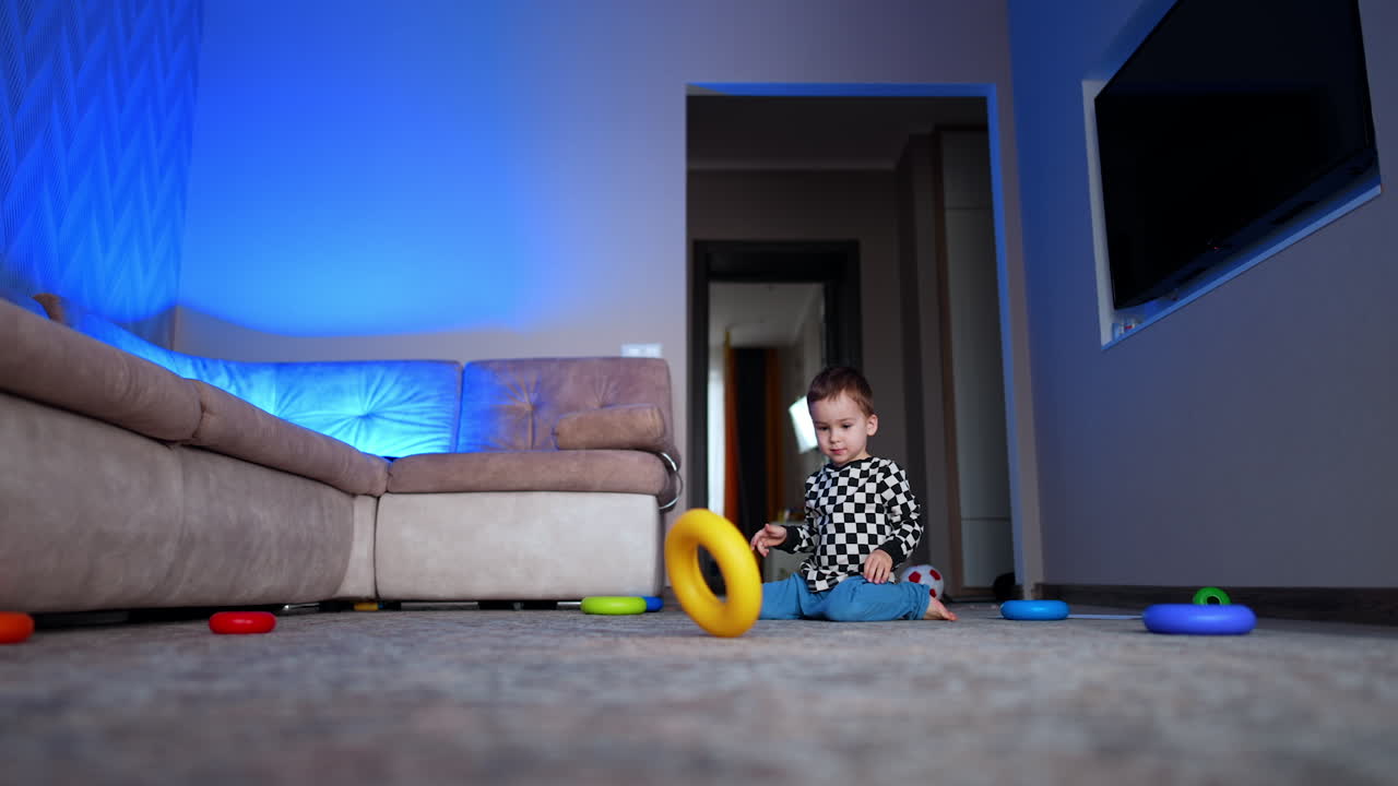 Cute child plays at home sitting on the floor. Caucasian baby rolls the pyramid rings cheerfully. Low angle view.