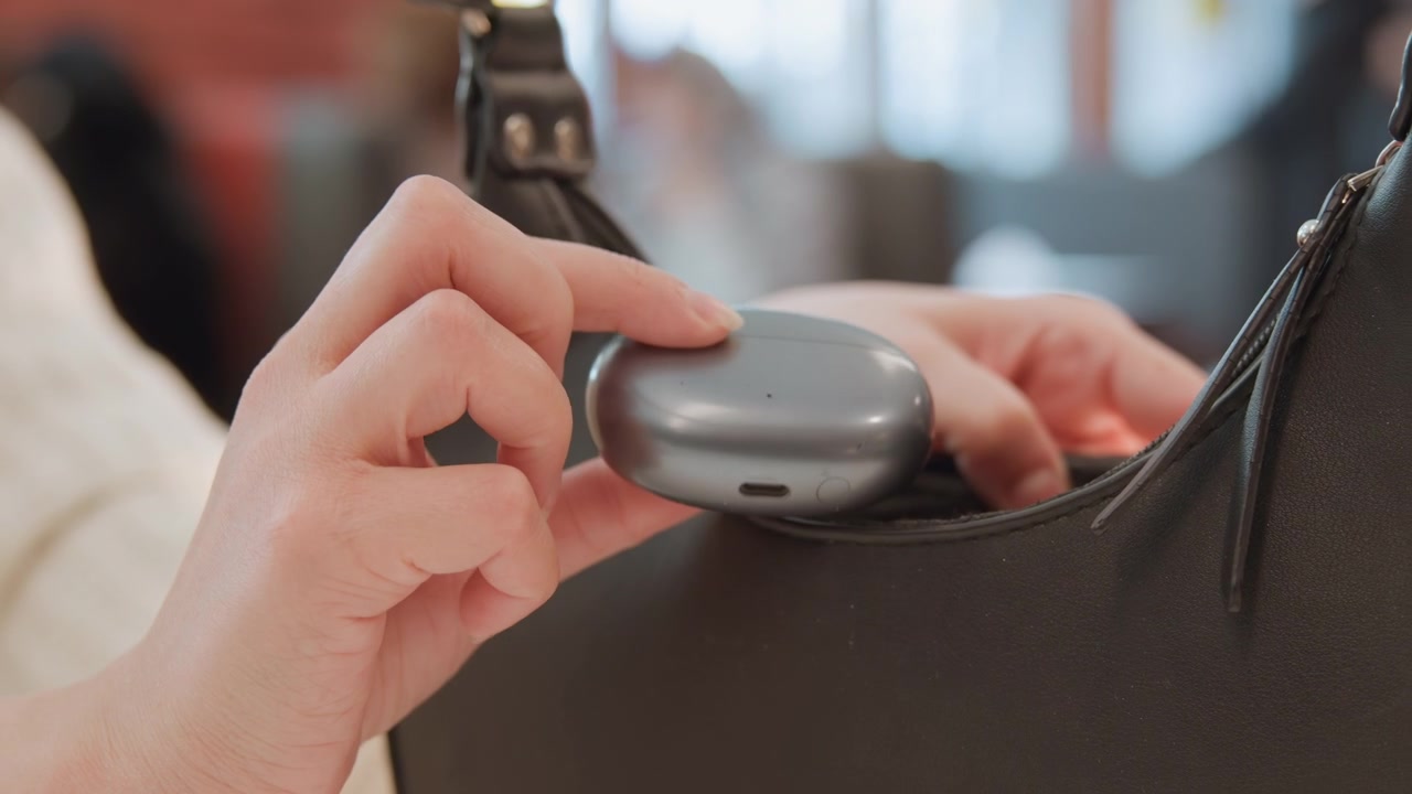 Azione ravvicinata della mano che mostra le dita posizionare la custodia degli auricolari wireless nella tasca di una borsa in pelle nera accanto a un bancone di negozio, con luce calda interna, sfocatura morbida e un contesto di shopping quotidiano.