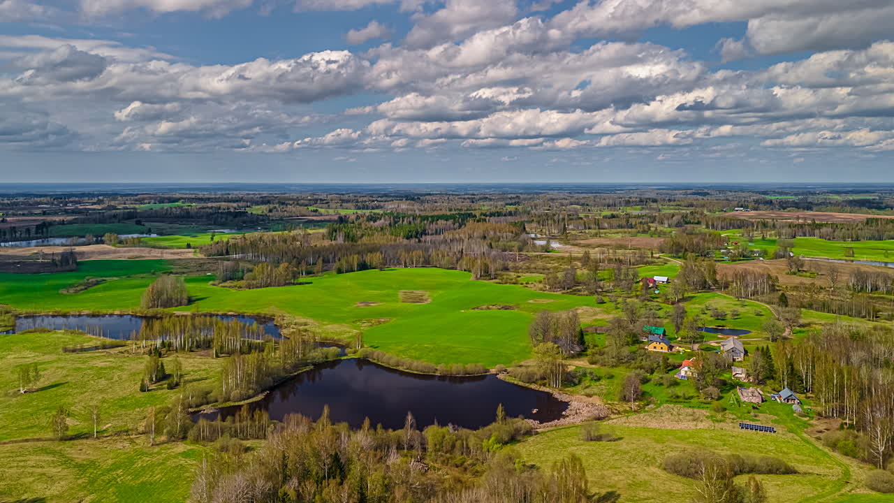 Farm village, meadows, fields, forests, and ponds in a rural landscape under clouds casting shadows, aerial time lapse