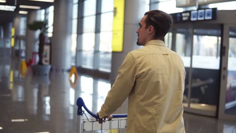 Brunette,-bearded-man-in-beige-shirt-walks-with-luggage-trolley-in-the-international-airport.-Walking-in-the-hurry,-rare