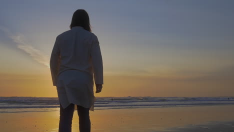 Man-in-kaftan-walks-on-beach-at-low-tide-in-golden-hour,-slow-motion