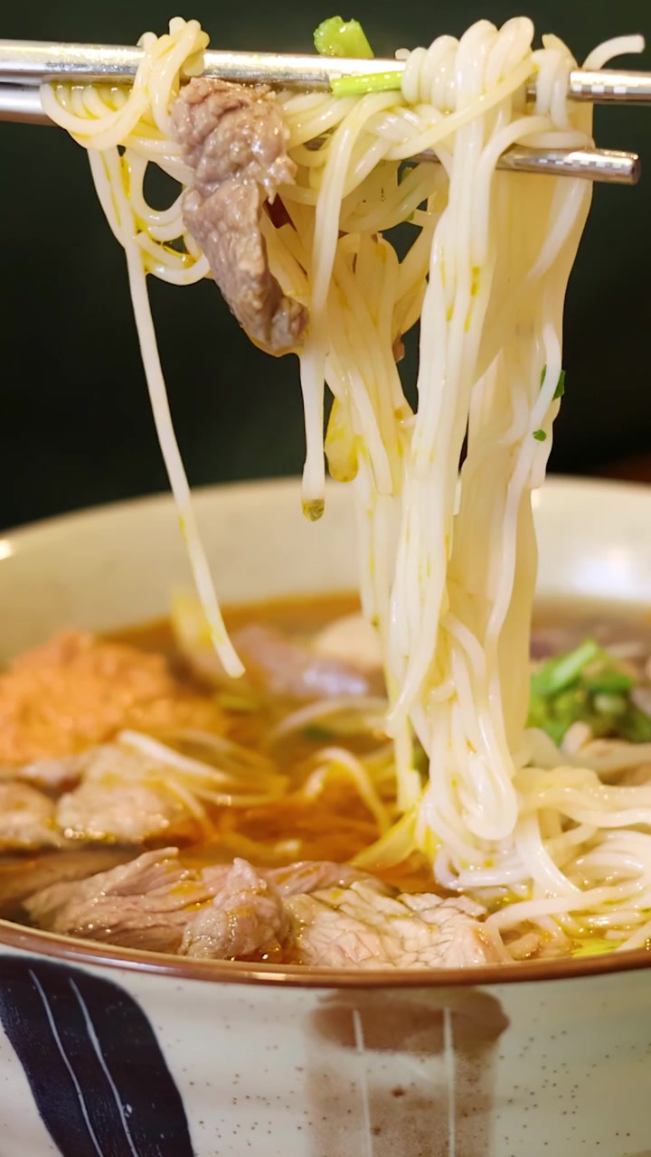 Un primo piano mostra bacchette di metallo che sollevano spaghetti di riso vermicelli da una ciotola di zuppa di manzo.