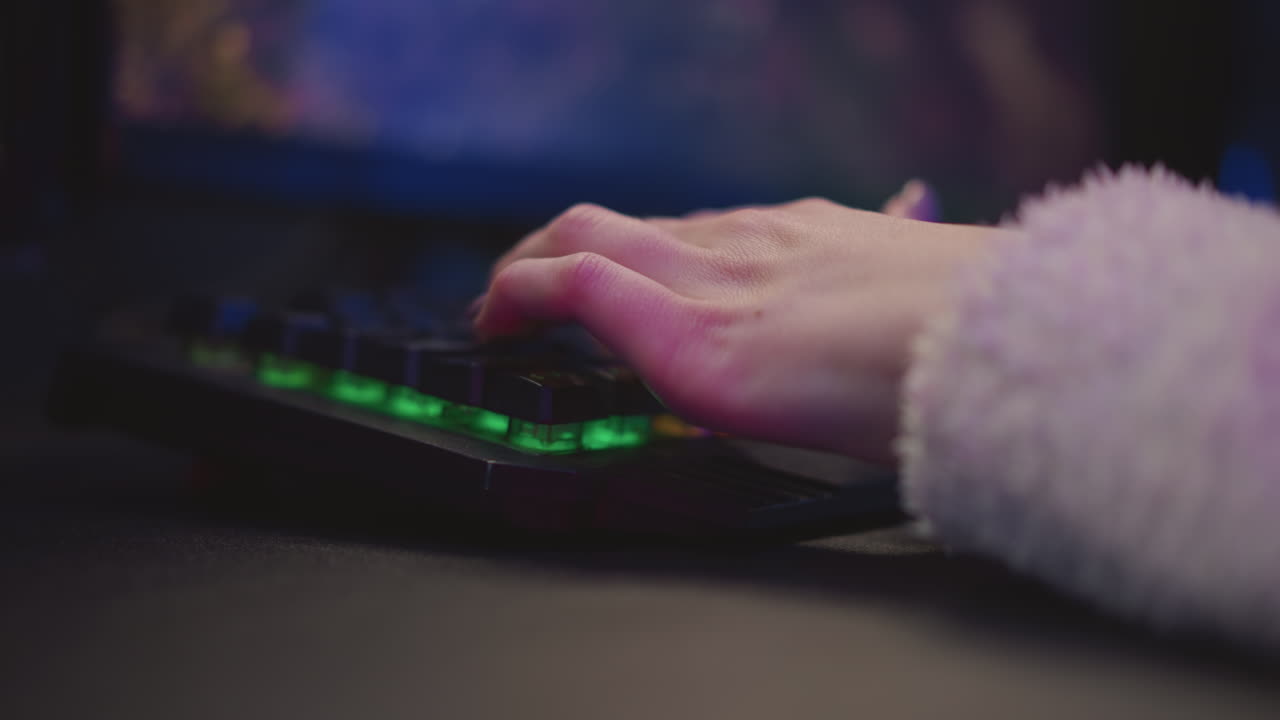 Close up side view of female gamer hand typing on illuminated keyboard with green backlight and fluffy sleeve visible, surrounded by soft ambient lighting and blurred screen in background