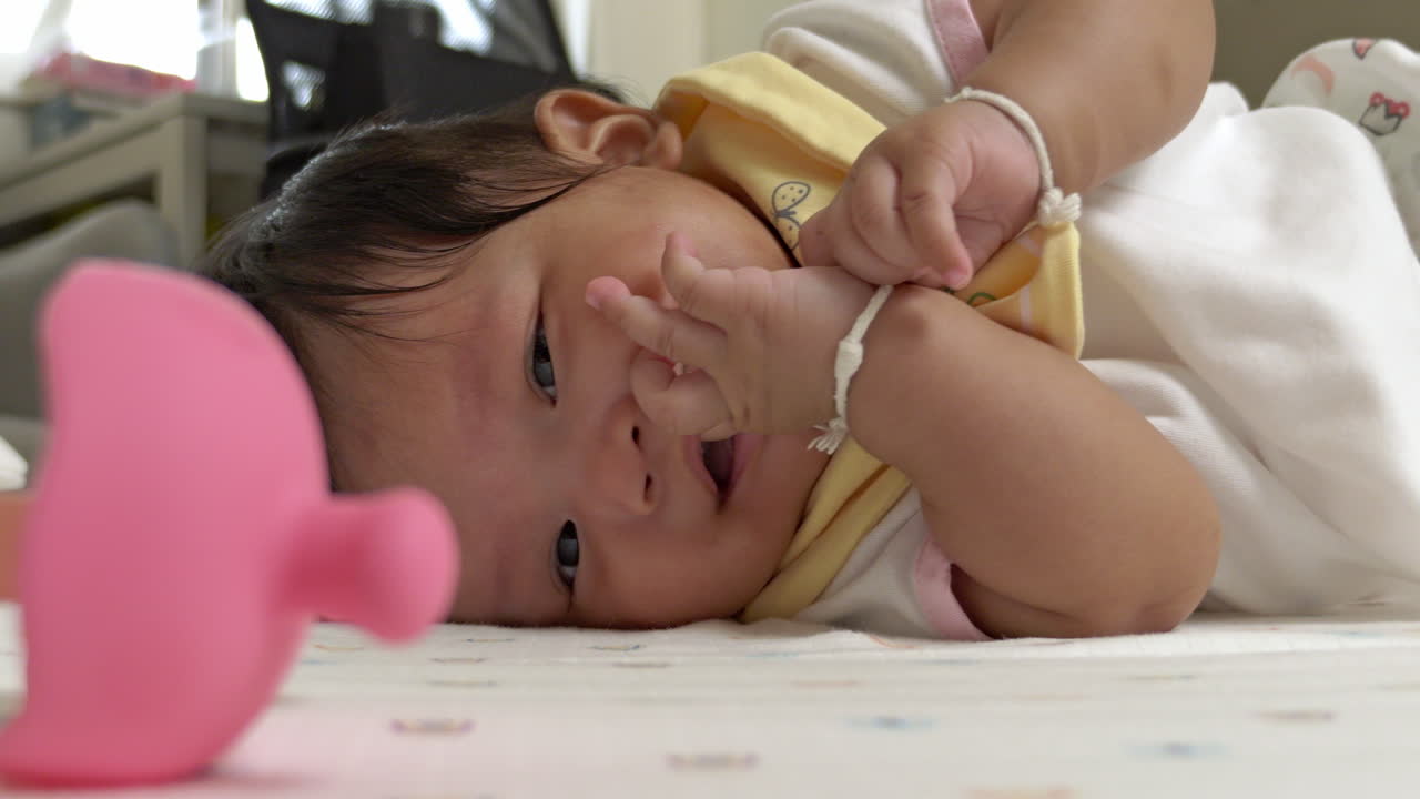 Charming video captures a baby girl happily sucking on her fingers while attempting to roll over on her mattress, showcasing early developmental milestones.