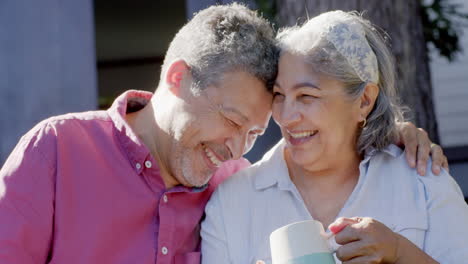 Happy-biracial-senior-couple-sitting,-embracing-and-drinking-tea-in-sunny-garden