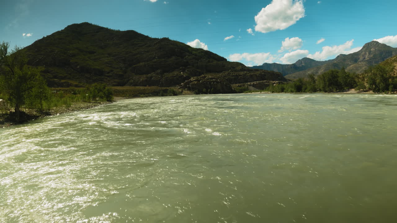 el río de montaña se precipita a lo largo de un amplio curso fpv. rápido arroyo de agua indomable brilla en la luz del sol de la bahía contra la gran montaña.