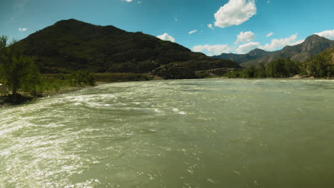 mountain river rushes along wide course fpv. fast untamed water stream glisten in bight sunlight against large mountain. fresh creek at highland