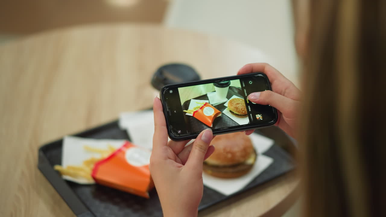dama vista desde atrás usando su teléfono inteligente para tomar una foto de su comida, que consiste en papas fritas, una hamburguesa y una taza de café, con los alimentos dispuestos cuidadosamente en una bandeja negra en una madera