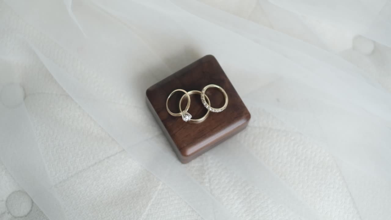 Evoke the romance of a wedding day with a close-up of engagement and wedding rings resting on a handcrafted wooden box, atop a plush white pillow, symbolizing lasting love and commitment.
