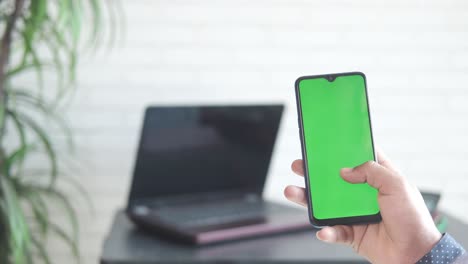 person holding a smartphone with a green screen, next to a laptop