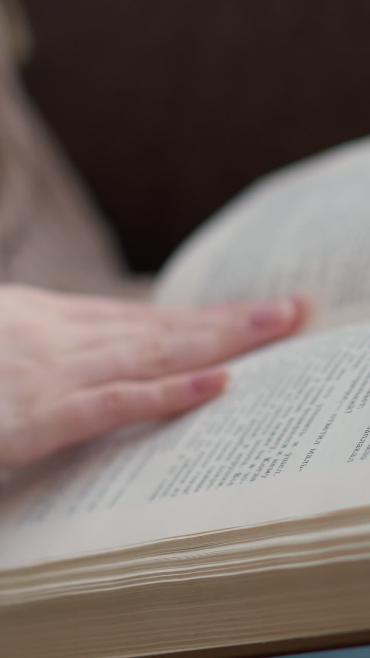 primer plano de una mano delicada con uñas bien cuidadas volviendo suavemente las páginas de un libro abierto, con ropa tejida, fondo ligeramente borroso