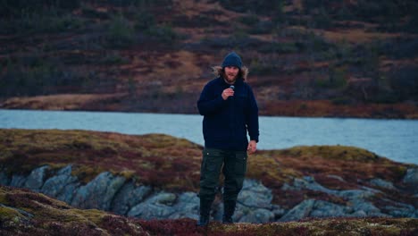 Man In Outdoor Gear Stands On Mossy Rock By The Reinsjoen Lake In Afjord, Norway, Talking On Lavalier Mic While Glancing Around. wide shot