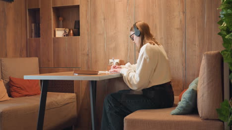 high school girl in white sweater wearing headset, gently dancing and writing at cozy wooden cafe booth, surrounded by warm lighting, green indoor plants, and soft cushions for a relaxed study vibe