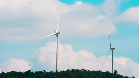 Dolly-out,-clean-renewable-energy-production,-futuristic-wind-turbines-spinning-slowly-in-the-middle-rural-countryside-generating-renewable-electric-energy-against-fluffy-clouds-and-cyan-sky