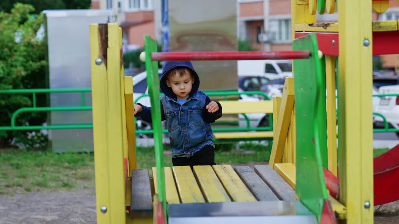 Sweet baby boy in a hood goes up by a ladder slowly. Careful kid manages to climb up and smiles happily.