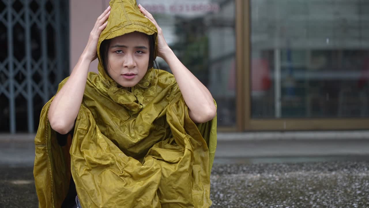 A young asian female in yellow hoody rain coat sit down waiting on the street side on the rainy day, stuck in the rain, self protection from, wet clothes stuck in the rain, wait for rain to stop