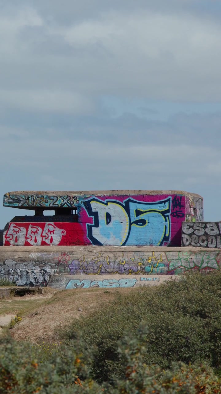 Colorful graffiti covers historic concrete bunker under daylight, static wide shot, coastal landscape visible