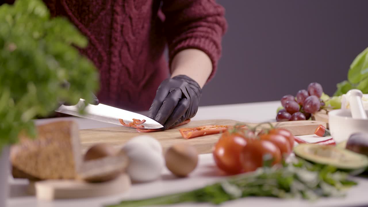 close-up of a chef chopping a red pepper on a wooden board. The sharp knife glides smoothly, revealing its crisp texture and seeds. Fresh ingredients like tomatoes, herbs, and avocado add depth