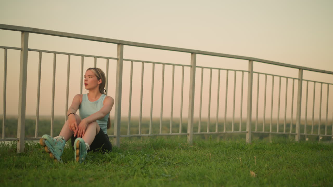 롤러 스케이트를 입은 젊은 여자가 철의 레일링 근처의 잔디에 앉아 무에 손을 고 깊은 숨을 들이마시며, 부드러운 저녁 빛과 흐릿한 배경의 초록색으로