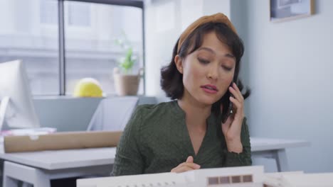 Biracial-businesswoman-talking-on-smartphone-and-building-model-alone-at-office