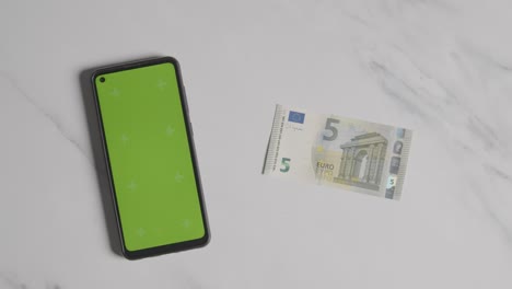 overhead currency shot of hand putting down us 5 euro note next to green screen mobile phone