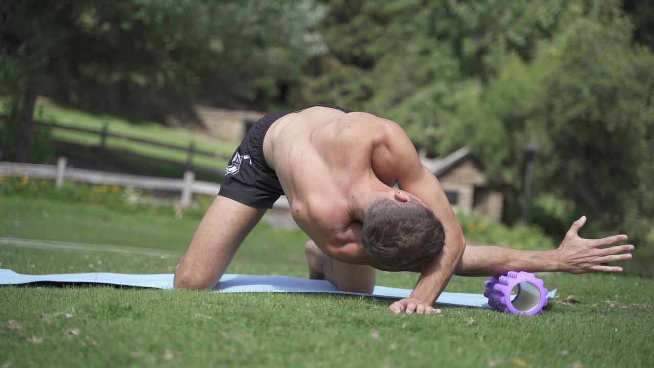 joven caucásico en forma atractiva masculina haciendo ejercicio al aire libre con rodillo de espuma hacia atrás con fondo de vegetación verde a cámara lenta
