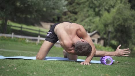 young caucasian fit attractive male exersicing outdoors foam roller back exercise with green vegetation background slow motion