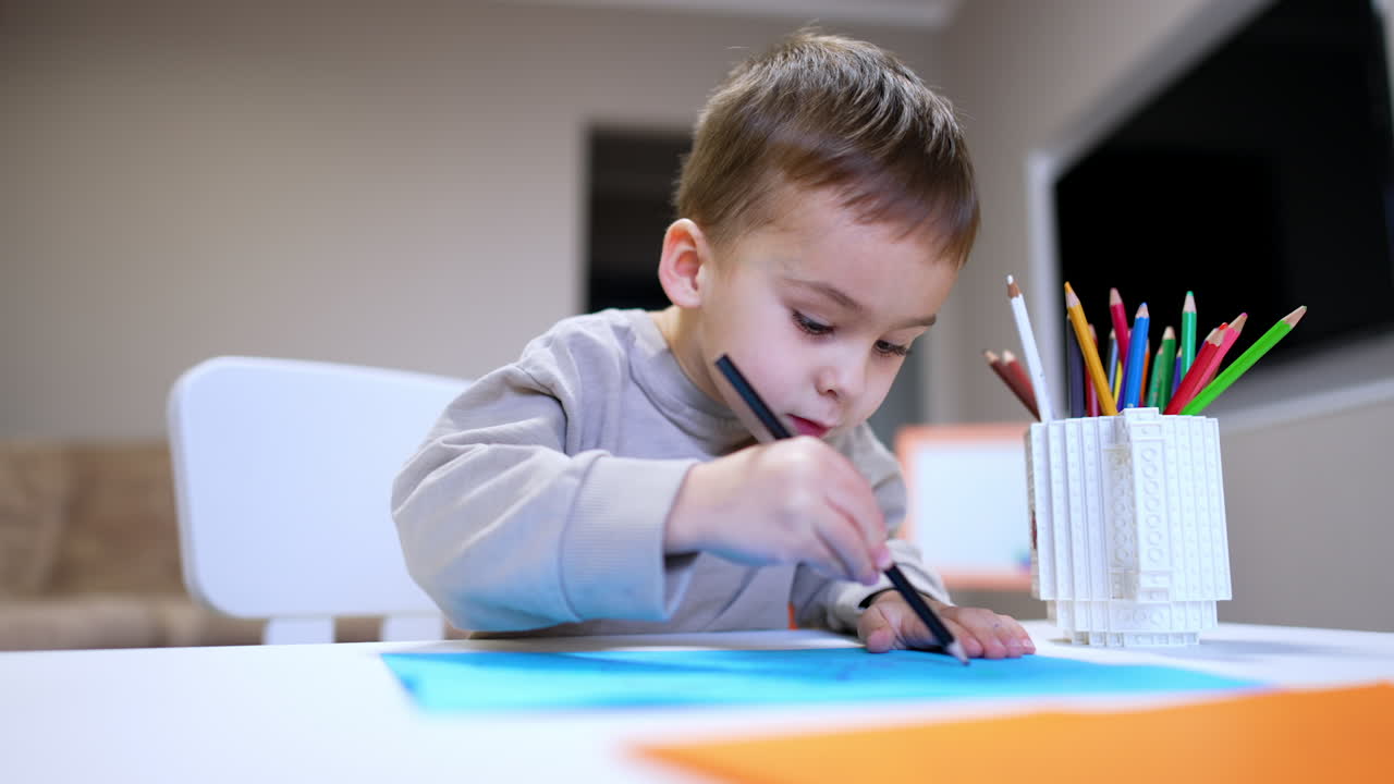 Lovely baby boy sits at desk talking. Beautiful toddler takes a pencil and starts to draw. Low angle view.