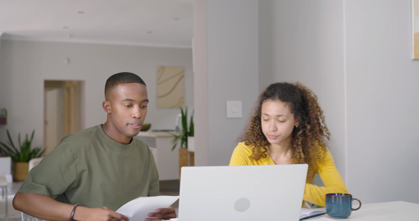 Homem e mulher afro-americanos consultando o laptop após uma ligação telefônica, sentados, espalhando papéis.