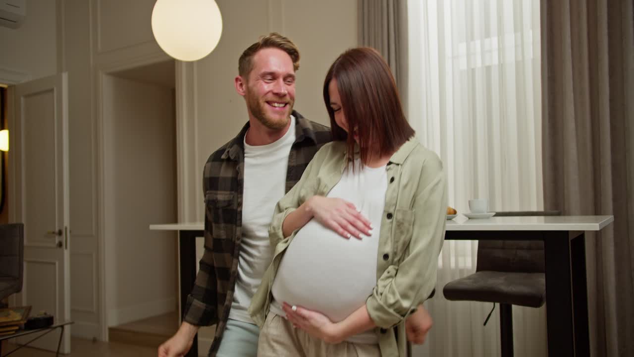 Happy blond man in a plaid shirt and white T-shirt dances with his pregnant brunette wife in a white T-shirt in a modern apartment during the day. Happy man dancing freestyle with his pregnant brunette girlfriend