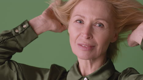 Vista-De-Cerca-De-Una-Mujer-Madura-Rubia-Con-Camisa-Verde-Sonriendo-A-La-Cámara-Tocando-Su-Cabello-Con-Viento-Sobre-Fondo-Verde