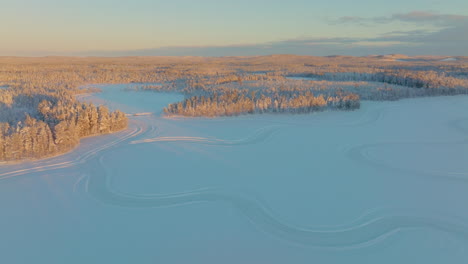 Golden-sunlit-Polar-circle-woodland-ice-lake-aerial-view-curving-tyre-tracks-under-glowing-sunrise-Norbotten-skyline