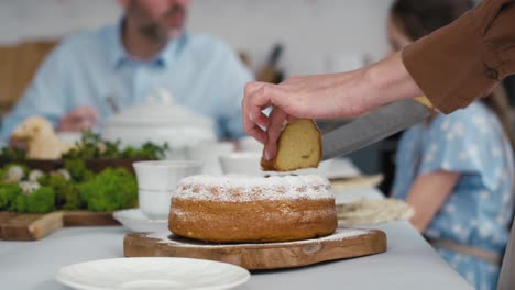 Unrecognizable-woman-cutting-easter-cake-and-passes-a-piece-to-the-family-in-the-background.