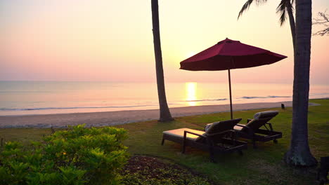 an empty pair of sun loungers under an open beach umbrella face the sunset as ocean waves roll in along the shore