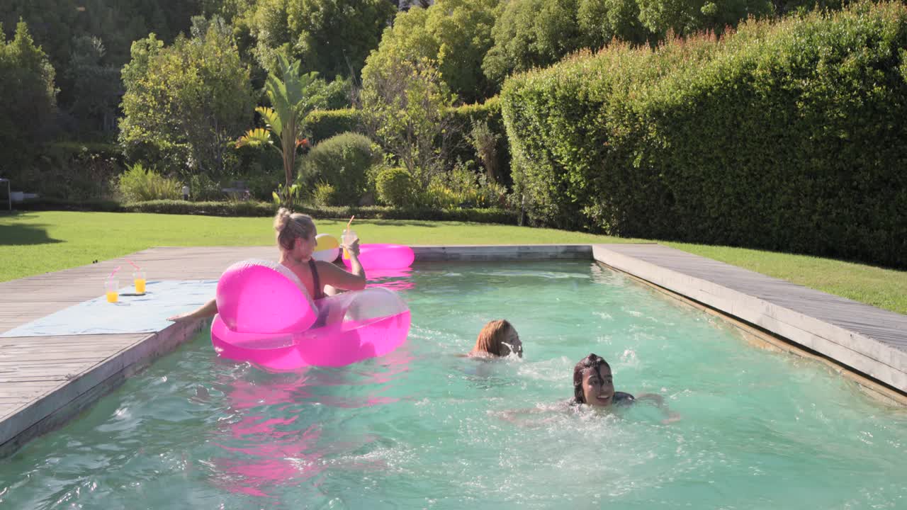 Female friends playing, jumping into pool, splashing woman on pink float holding drink, copy space