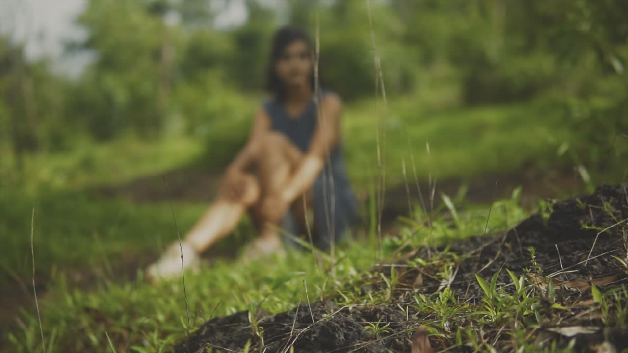 Close up fixed shot of earth and grass seedlings on the side of a gentle hill, with female south asian model sitting in the grass blurred in the background